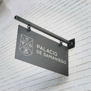 Black engraved metal hanging sign with silver logo engraving on a powder coated black wall bracket.